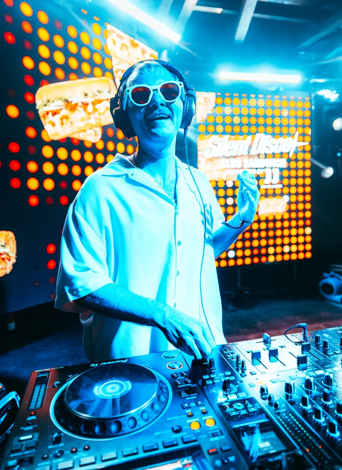 TITAN smiling behind the decks with one hand raised under blue stage lights