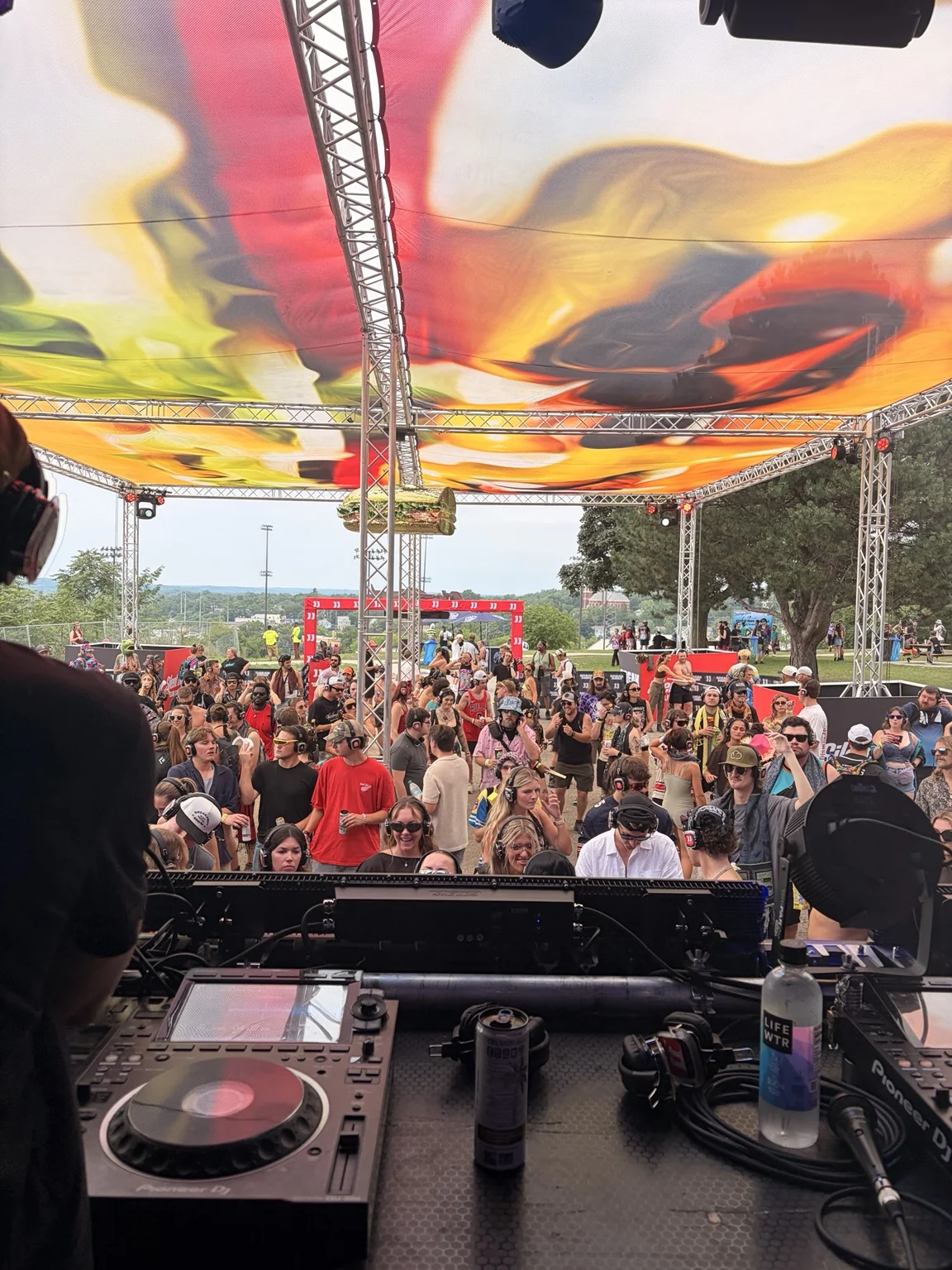 Daytime festival crowd beneath a colorful canopy, seen from behind the DJ booth